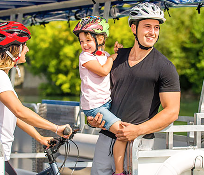 Un couple et un enfant profite d'une balade en navette fluviale sur le Saint-Laurent à Montréal. La femme tient un vélo et porte un casque sur la tête,  l'homme porte l'enfant dans ses bras.