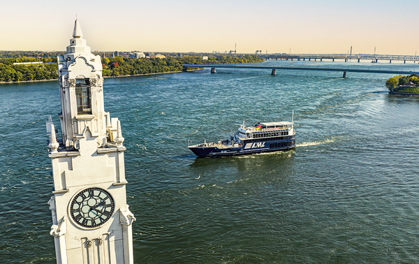 The AML Cavalier Maxim ship sailing on the Saint Lawrence River with the Montreal Clock Tower in the foreground and the Saint Helen's Island and the Samuel de Champlain Bridge in the background with a golden light.