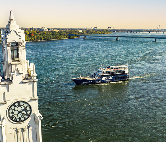 The AML Cavalier Maxim ship sailing on the Saint Lawrence River with the Montreal Clock Tower in the foreground and the Saint Helen's Island and the Samuel de Champlain Bridge in the background with a golden light.