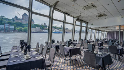 Salle intérieure avec des tables dressées devant une fenêtre donnant vue sur le fleuve Saint-Laurent, le Château Frontenac et le cap Diamant à Québec en arrière plan 