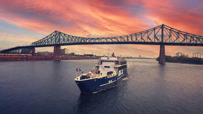 Un bateau de croisière bleu marqué "AML" navigue sur un fleuve au coucher du soleil, avec le pont Jacques-Cartier en arrière-plan sous un ciel spectaculaire aux teintes roses et oranges.