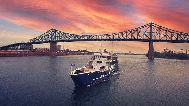 Un bateau de croisière bleu marqué "AML" navigue sur un fleuve au coucher du soleil, avec le pont Jacques-Cartier en arrière-plan sous un ciel spectaculaire aux teintes roses et oranges.