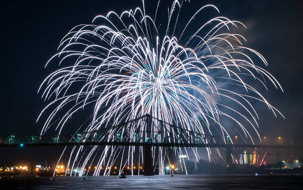 Un feu d'artifice spectaculaire éclate dans le ciel nocturne au-dessus d'un pont illuminé, avec des reflets sur l'eau et des lumières urbaines en arrière-plan.