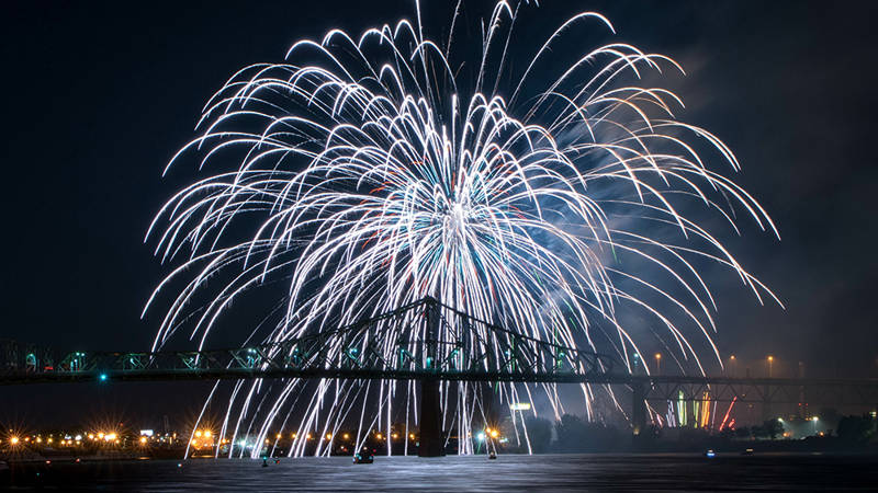 Un feu d'artifice spectaculaire éclate dans le ciel nocturne au-dessus d'un pont illuminé, avec des reflets sur l'eau et des lumières urbaines en arrière-plan.