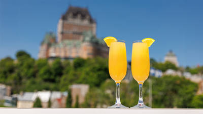 Deux cocktails mimosas, garnis de tranches d'orange sur le bord du verre, sont placés au premier plan. En arrière-plan flou, on distingue un bâtiment historique orné, avec un toit vert et un ciel bleu au-dessus.