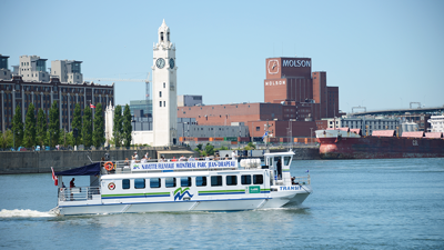 Une navette fluviale naviguant sur le fleuve Saint-Laurent avec le fleuve au premier plan et la tour de l'horloge et le bâtiment de la brasserie Molson en arrière plan.