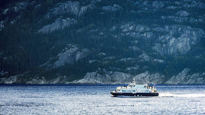 Un bateau de passagers navigue sur un lac calme, avec des collines rocheuses et boisées en arrière-plan. Le soleil, partiellement voilé par des nuages, se trouve au-dessus des collines, ajoutant une touche douce à l'atmosphère.