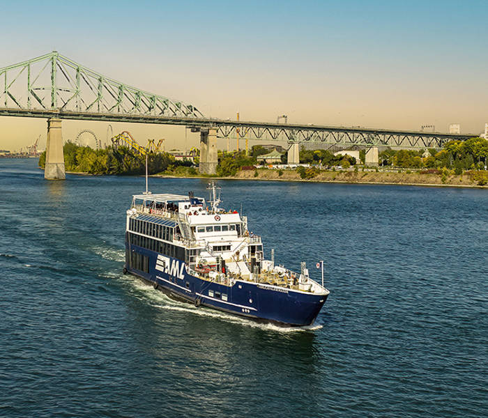 Un bateau de croisière navigue sur une rivière par une journée ensoleillée, avec le pont Jacques-Cartier en arrière-plan et des arbres bordant la rive.