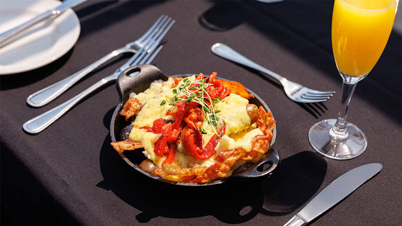 Un plat chaud servi dans une petite poêle en fonte, garni de légumes grillés, de fromage fondu et de pousses fraîches, accompagné d'un verre de mimosa sur une table élégamment dressée.