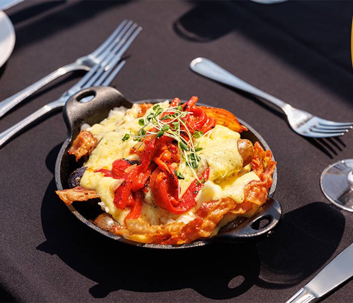 Un plat chaud servi dans une petite poêle en fonte, garni de légumes grillés, de fromage fondu et de pousses fraîches, accompagné d'un verre de mimosa sur une table élégamment dressée.