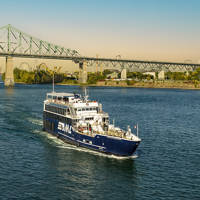 Un bateau de croisière bleu navigue sur une rivière par une journée ensoleillée, avec le pont Jacques-Cartier et des arbres en arrière-plan.