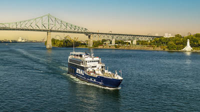 Un bateau de croisière bleu navigue sur un fleuve par une journée ensoleillée, avec le pont Jacques-Cartier et une rive verdoyante en arrière-plan.