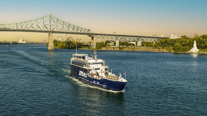 Un bateau de croisière bleu navigue sur un fleuve par une journée ensoleillée, avec le pont Jacques-Cartier et une rive verdoyante en arrière-plan.