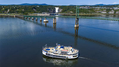 Un ferry blanc à plusieurs ponts navigue sur une large rivière, avec un pont suspendu vert visible au milieu de la scène. Au-delà du pont, une cascade dévale une falaise verdoyante. Un paysage vallonné et boisé borde la rivière, le tout sous un ciel bleu clair.