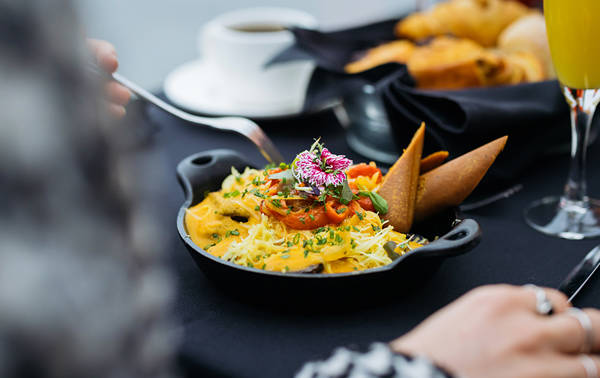 Un plat élégant et coloré garni de fleurs comestibles, servi dans une petite poêle noire sur une table avec des viennoiseries, une tasse de café et un verre de jus.