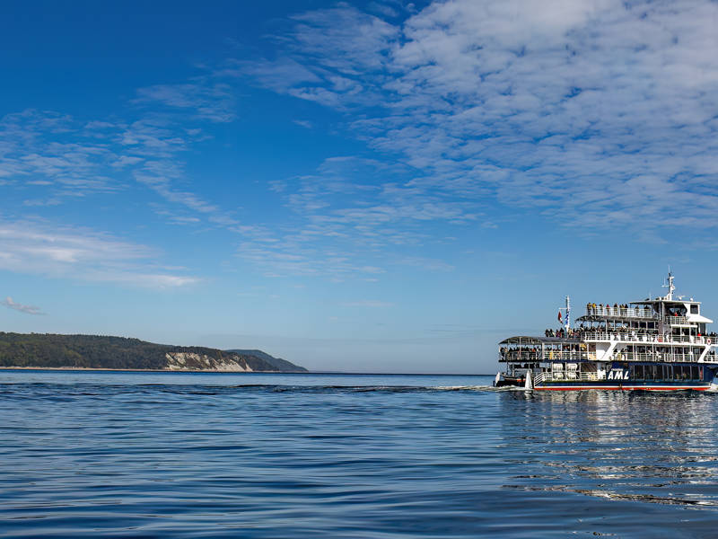 Un ferry navigue sur une étendue d'eau paisible sous un coucher de soleil doré. Le ciel est partiellement nuageux, et des collines recouvertes d'une dense forêt verte encadrent la scène en arrière-plan. Le ferry laisse derrière lui un sillage doux qui se dissipe lentement.