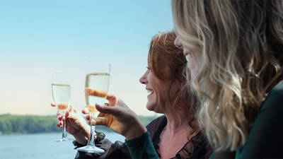 Deux femmes sourient et trinquent avec des coupes de champagne, profitant d'une journée ensoleillée avec une vue sur l'eau et les arbres en arrière-plan.