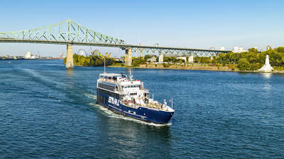 Un bateau de tourisme bleu navigue sur une large rivière sous un pont en acier vert. La scène ensoleillée révèle l'horizon d'une ville au loin et des arbres luxuriants bordant les rives, le tout sous un ciel bleu clair.