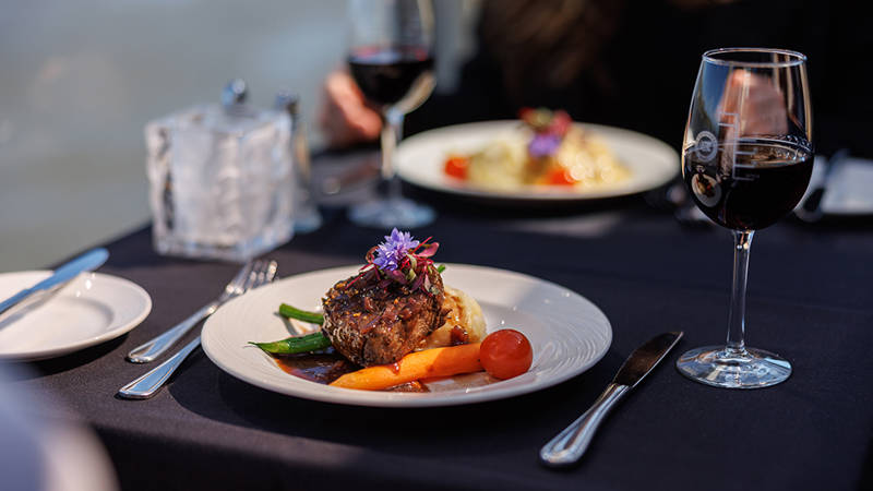 Une assiette élégamment dressée contenant une pièce de viande garnie de légumes et de fleurs comestibles, accompagnée de verres de vin rouge sur une table avec une nappe sombre.