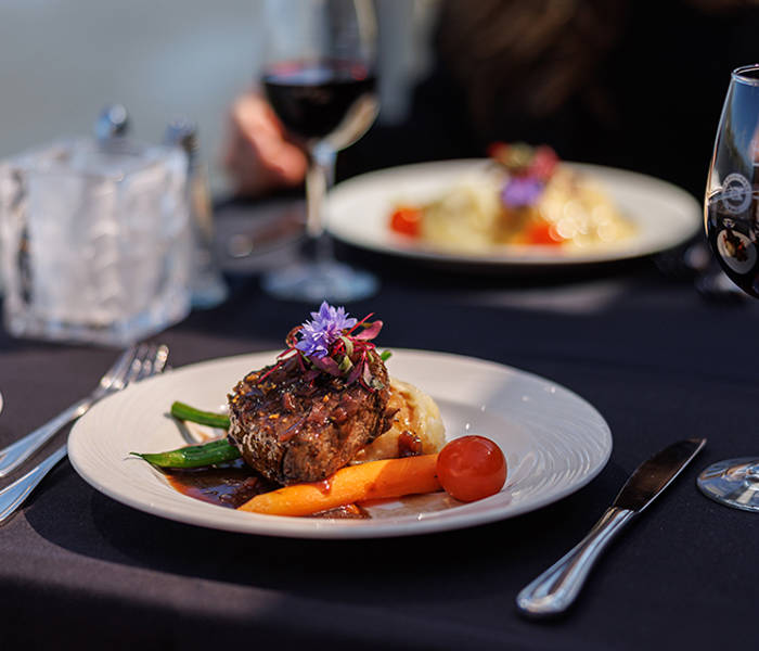 Une assiette élégamment dressée contenant une pièce de viande garnie de légumes et de fleurs comestibles, accompagnée de verres de vin rouge sur une table avec une nappe sombre.