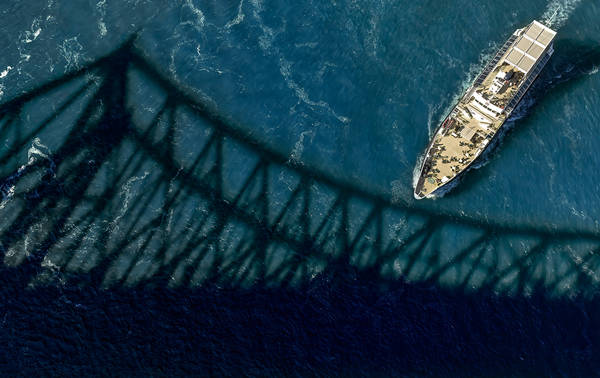 Vue aérienne d'un bateau naviguant sur une rivière bleue, avec l'ombre imposante d'un pont projetée sur l'eau.