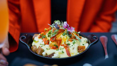 Un bol de gratin de pommes de terre au fromage, garni de ciboulette hachée, de poivrons rouges rôtis et de fleurs comestibles. Il est servi dans un bol noir, posé sur une table à côté d'une fourchette et d'un couteau, avec une veste orange visible en arrière-plan.