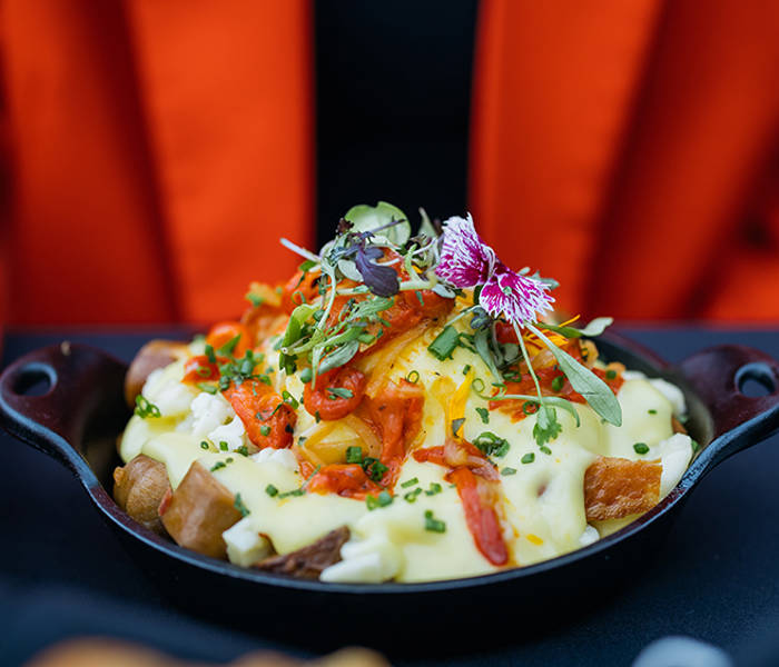 Un bol de gratin de pommes de terre au fromage, garni de ciboulette hachée, de poivrons rouges rôtis et de fleurs comestibles. Il est servi dans un bol noir, posé sur une table à côté d'une fourchette et d'un couteau, avec une veste orange visible en arrière-plan.