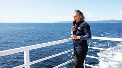 A naturalist stands on a boat's deck holding a microphone, overlooking the vast ocean under a clear blue sky. Distant landforms are visible across the water. The person wears a dark outfit and appears to be speaking or singing.