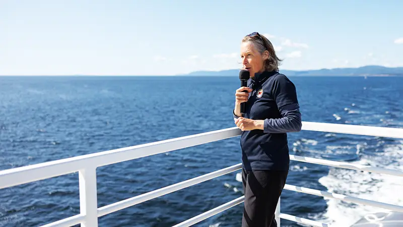 A naturalist stands on a boat's deck holding a microphone, overlooking the vast ocean under a clear blue sky. Distant landforms are visible across the water. The person wears a dark outfit and appears to be speaking or singing.
