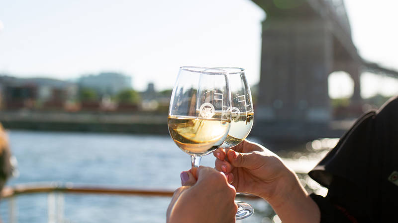 Deux personnes trinquent avec des verres de vin blanc sur un bateau, avec une rivière et un pont en arrière-plan sous un ciel ensoleillé.