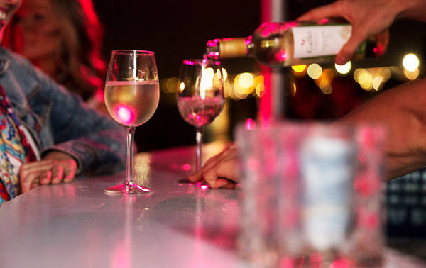 Un serveur verse du vin blanc dans des verres sur un bar sous une lumière tamisée rose, avec des clients souriants en arrière-plan.