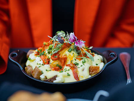 Un plat gourmand composé de pommes de terre rôties, nappées de sauce hollandaise, garnies de légumes, herbes fraîches et fleurs comestibles, servi dans une poêle en fonte.