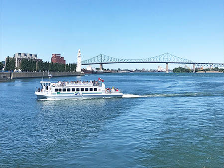 Une navette fluviale traverse le fleuve sous un ciel bleu, avec la tour de l'Horloge, le pont Jacques-Cartier et des bâtiments de Montréal visibles en arrière-plan.