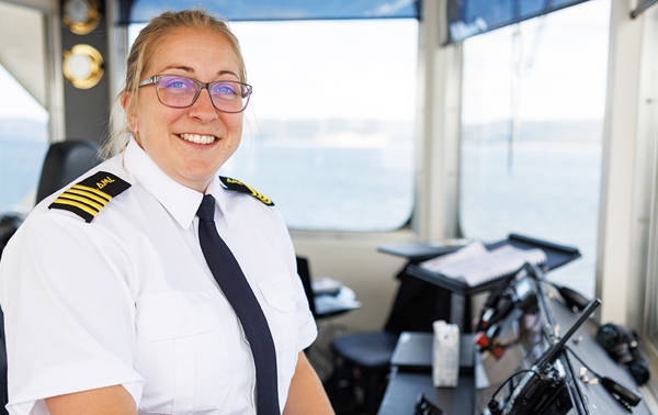 Une personne portant un uniforme de capitaine, comprenant une chemise blanche avec des épaulettes et une cravate noire, sourit avec une sucette dans la bouche. Elle se trouve dans la salle de contrôle d'un navire entourée d'équipements de navigation. Une grande fenêtre offre une vue sur une étendue d'eau.