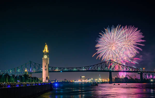 Un feu d'artifice coloré éclate dans le ciel nocturne près du pont Jacques-Cartier et de la tour de l'Horloge, avec des reflets sur le fleuve et une foule observant depuis le quai.