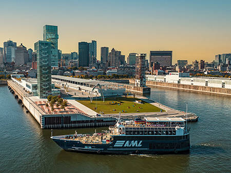 Un bateau de croisière bleu avec le logo "AML" navigue près d'un quai moderne avec des espaces verts, entouré par les gratte-ciels d'une ville sous un ciel clair et ensoleillé.