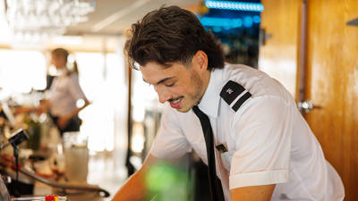 Un barman en uniforme blanc avec une cravate noire prépare une boisson dans un bar lumineux et moderne, avec une ambiance professionnelle et accueillante.