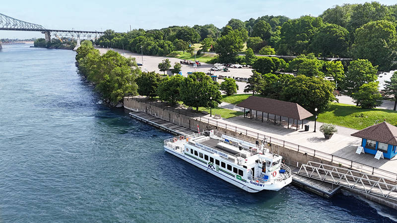 Une vue aérienne d'un bateau de croisière amarré le long d'un quai bordé d'arbres, avec un parc verdoyant en arrière-plan et un pont visible au loin.