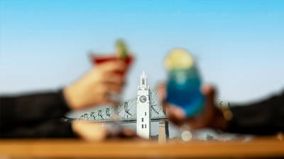 Deux personnes trinquent avec des cocktails colorés (un rouge et un bleu) en arrière-plan flouté d'une tour blanche avec une horloge et un pont.