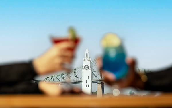 Deux personnes trinquent avec des cocktails colorés (un rouge et un bleu) en arrière-plan flouté d'une tour blanche avec une horloge et un pont.