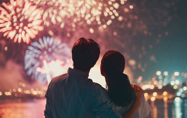 Un couple regarde un feu d'artifice coloré illuminant le ciel nocturne, avec des reflets scintillants sur l'eau et des lumières urbaines en arrière-plan.