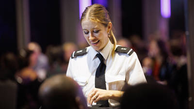 Une serveuse souriante en uniforme blanc et cravate noire interagit avec des clients dans un cadre élégant et animé, avec des lumières tamisées en arrière-plan.