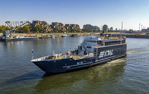 Un bateau de croisière noir avec l'inscription "AML" naviguant sur une rivière. En arrière-plan, un complexe résidentiel distinctif, un pont et des bâtiments urbains sous un ciel bleu dégagé.