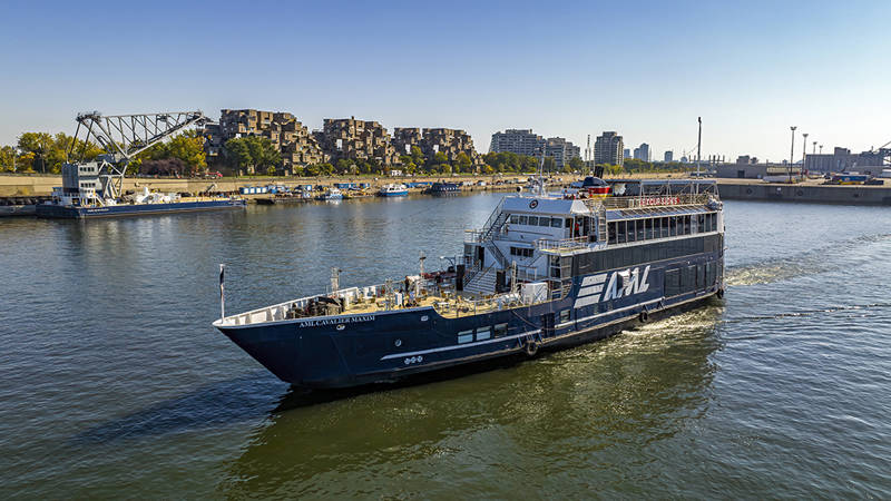 Un bateau de croisière noir avec l'inscription "AML" naviguant sur une rivière. En arrière-plan, un complexe résidentiel distinctif, un pont et des bâtiments urbains sous un ciel bleu dégagé.