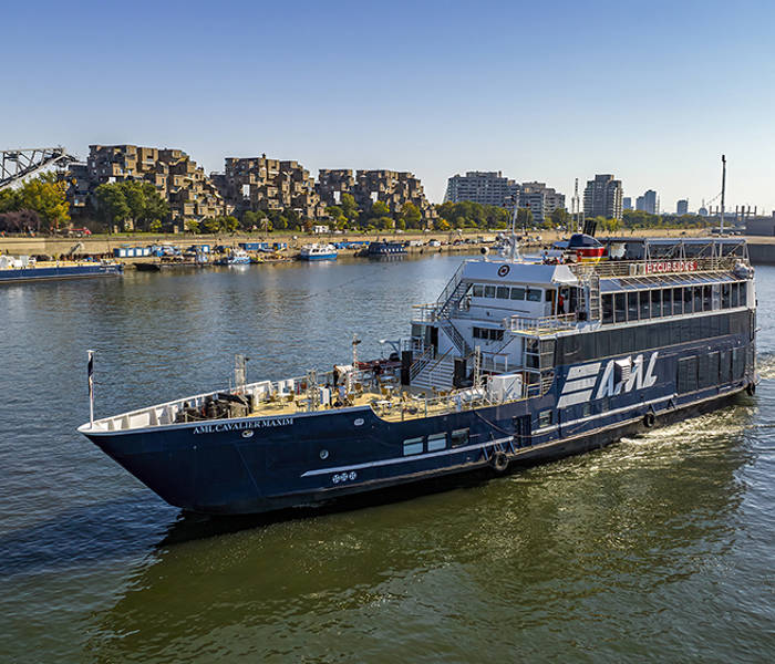 Un bateau de croisière noir avec l'inscription "AML" naviguant sur une rivière. En arrière-plan, un complexe résidentiel distinctif, un pont et des bâtiments urbains sous un ciel bleu dégagé.