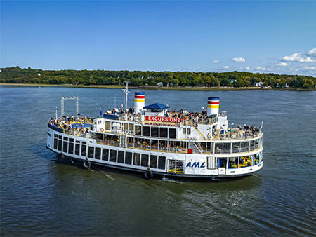 Un grand ferry blanc avec deux cheminées colorées navigue sur une large rivière. Il est rempli de passagers. En arrière-plan, on peut voir des terres boisées et un ciel bleu partiellement nuageux.