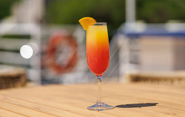 Un verre de cocktail coloré rouge et jaune, garni d'une tranche d'orange, posé sur une table en bois avec un arrière-plan flou comprenant un bateau et des éléments nautiques.