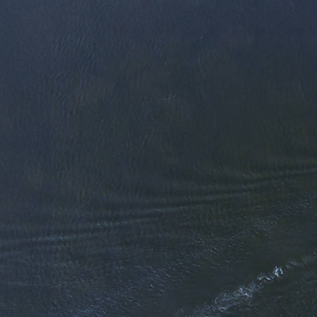 Aerial view of the Louis Jolliet on a vast expanse of the blue waters of the St. Lawrence. The boat moves forward, leaving gentle ripples behind. It has multiple decks with visible structures and passengers on board.