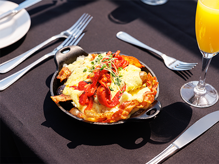 Cassolette gourmande comprise dans le brunch avec œufs brouillés, pommes de terre grelots, poivrons rôtis, fromage en grains, bacon, saucisses et sauce hollandaise accompagnée d'un verre de mimosa 