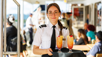 Une serveuse souriante en uniforme tenant un plateau avec deux cocktails de couleur orangée posés dessus, le tout sur le navire AML Cavalier Maxim à Montréal. 